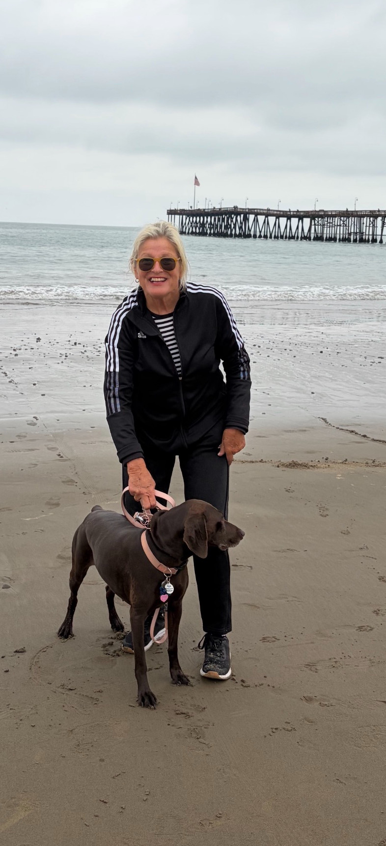 Jann Meyers smiling with a brown dog on the beach near a pier.